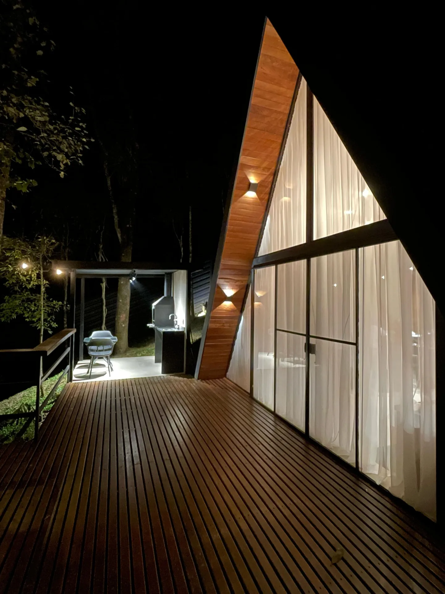Fachada A-frame de vidro iluminada à noite, vista do deck